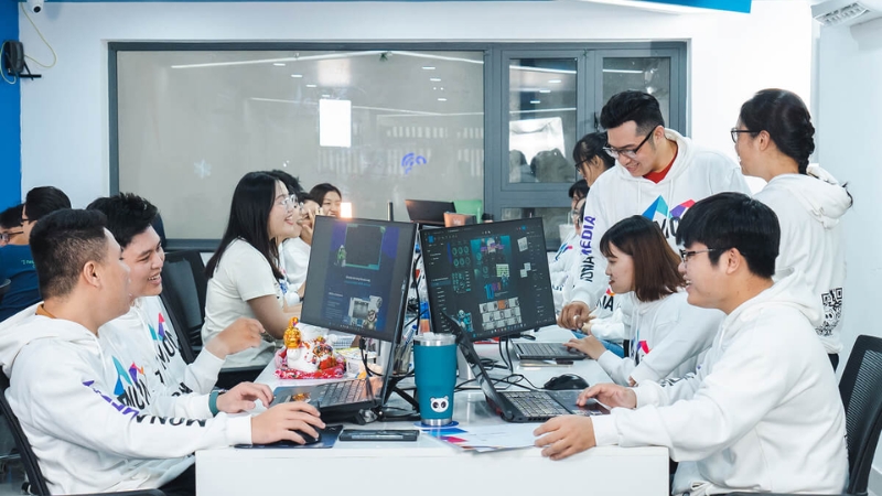 Đội ngũ Mona Media mặc áo hoodie trắng in logo làm việc trên máy tính tại văn phòng công ty SEO TP.HCM