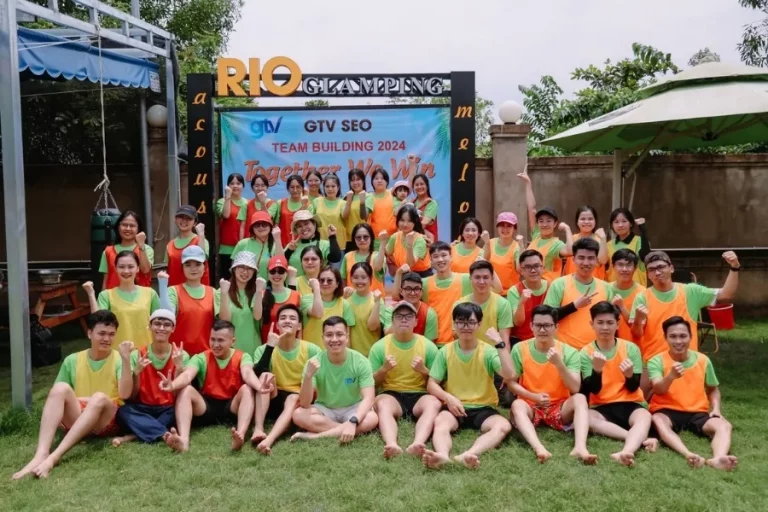 Đội ngũ GTV SEO mặc áo nhiều màu theo nhóm, ngồi trên bãi cỏ dưới băng rôn GTV SEO Team Building 2024